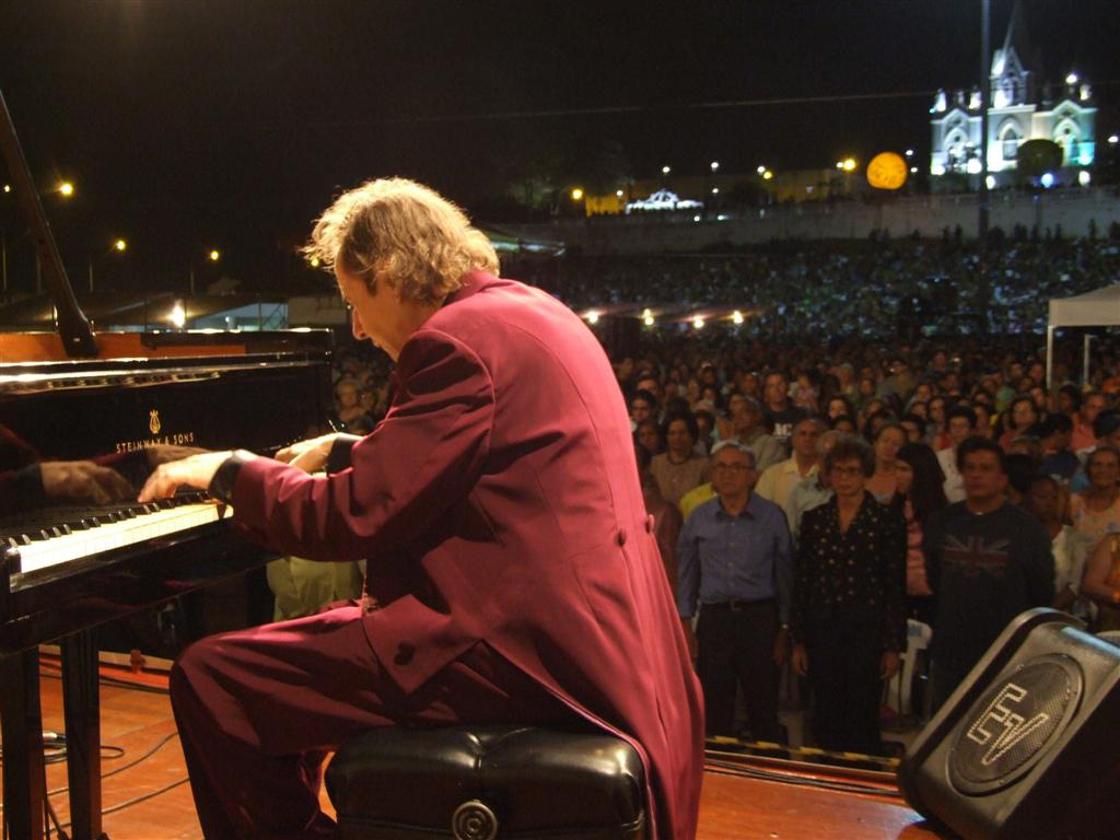 Pianista em primeiro plano toca para plateia numerosa de pé ao fundo